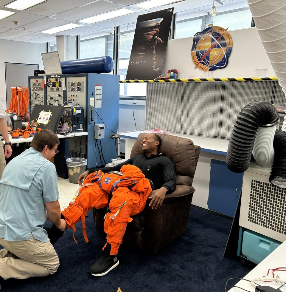 an astronaut sitting down on a chair with a technician trying to put on a shoe and a bulky spacesuit legging. the astronaut is laughing