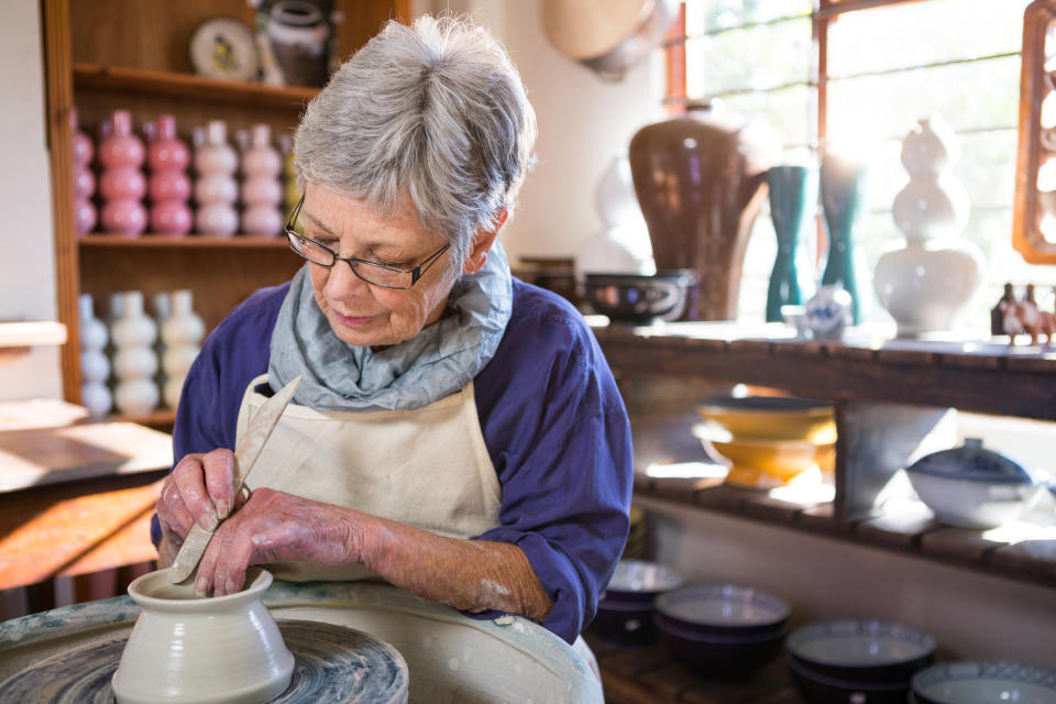 Gray-haired person making a clay pot in a shop with many materials.