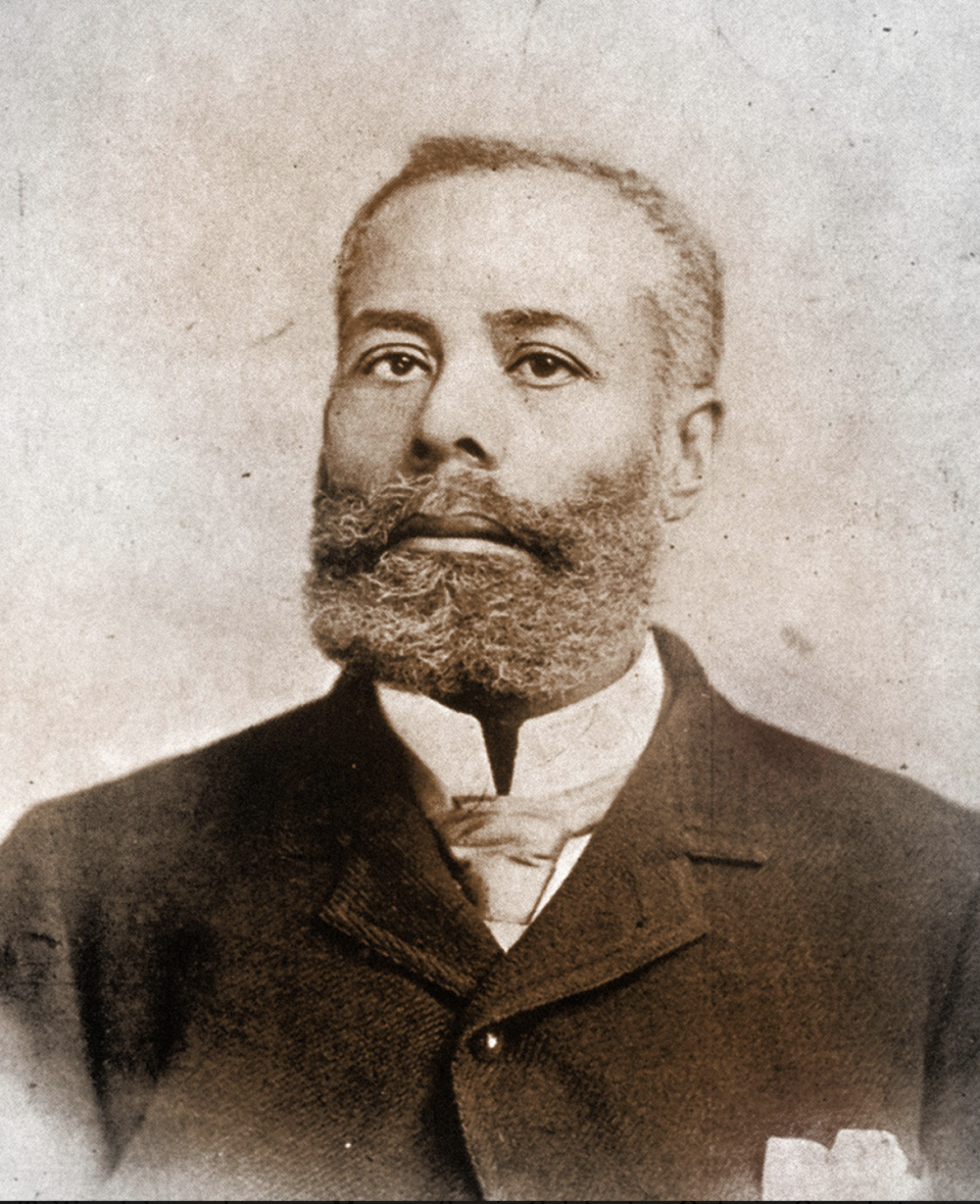 elijah mccoy looks at the camera, he wears a jacket over a collared shirt and tie and has a full beard