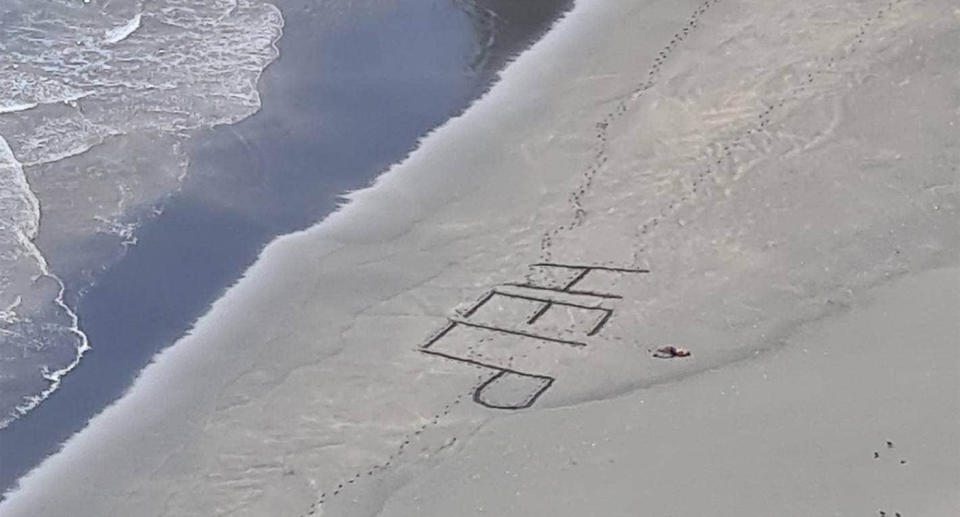 A message in the sand reads 'HELP' as a seriously injured surfer lies nearby, exhausted.