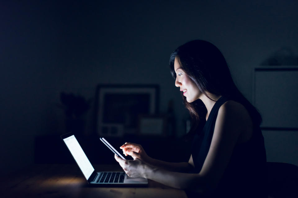 Busy young woman text messaging on smartphone while working on laptop till late in home office