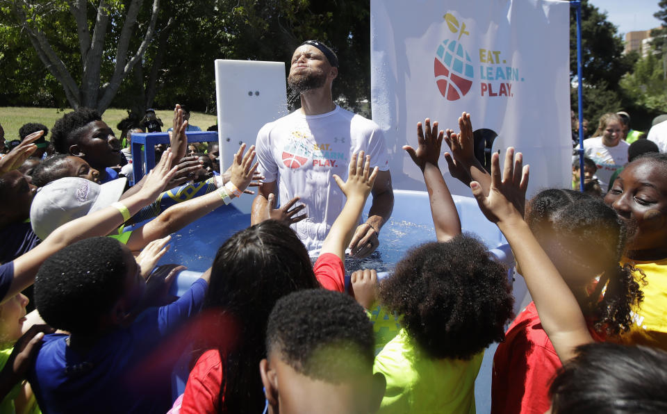 FILE - Golden State Warriors basketball player Stephen Curry, center, is cheered by kids after being dunked in a tank as part of the launching of the Eat. Learn. Play. Foundation in Oakland Calif., July 18, 2019. Curry, his wife Ayesha Curry, and partners are expanding the reach of their Eat.Learn.Play. Foundation, established in 2019 to support youth in Oakland, the Bay Area and beyond, while striving to improve lives of families nationwide. They are generating $50 million in additional funding to assist the Oakland Unified School District. (AP Photo/Jeff Chiu, File)