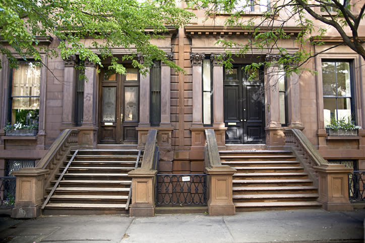 Two brownstone stoop entrances side by side, each with steps leading up to a front door, surrounded by trees and railings