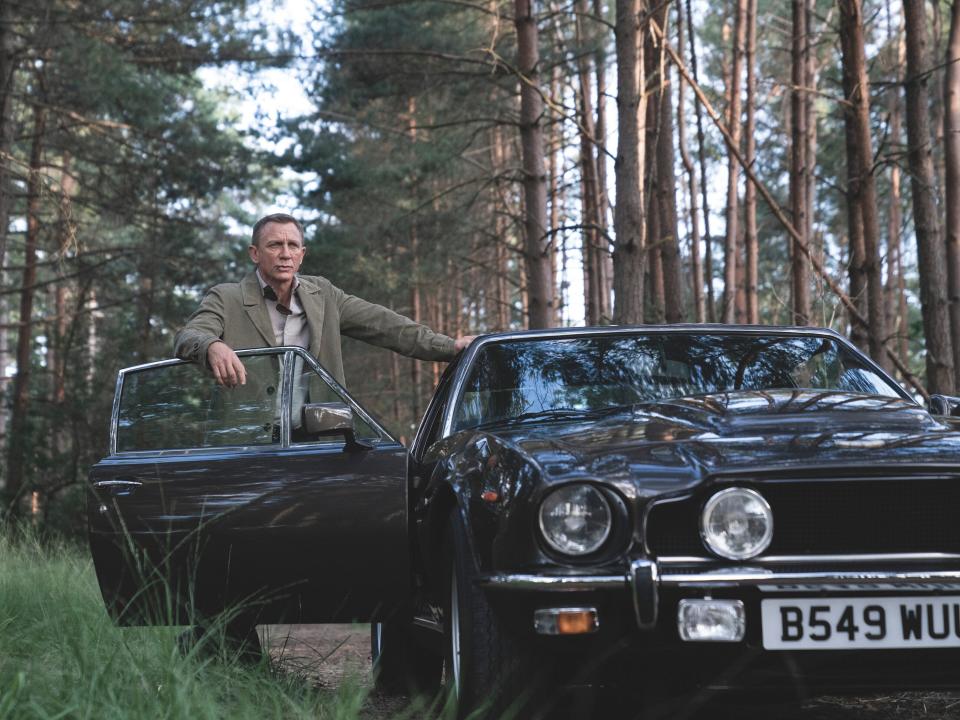 Daniel Craig standing in a forest beside an Aston Martin V8.