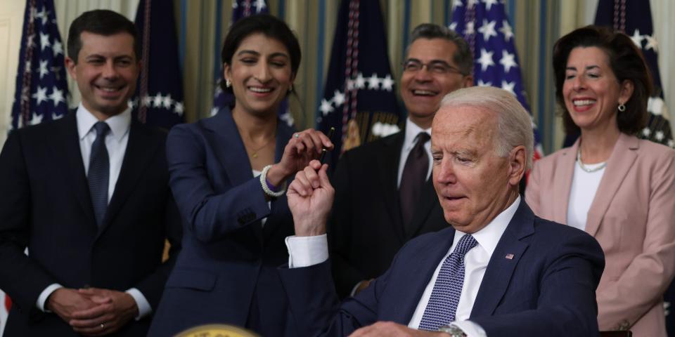 Lina Khan hands a pen to President Joe Biden, sitting at a desk with the presidential seal, with three other Cabinet officials behind him.