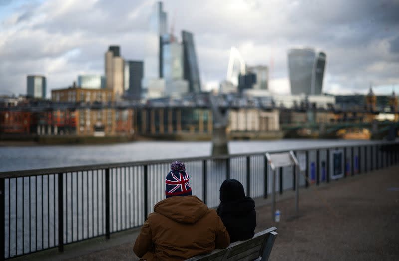 People sit alongside the bank of the River Thames with the City of London financial district in the background, in London