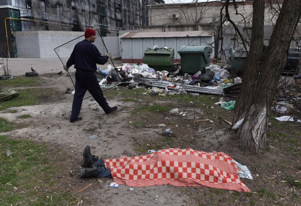 A local resident carries a broken window past the body of a person covered with a red-and-white checkered cloth.