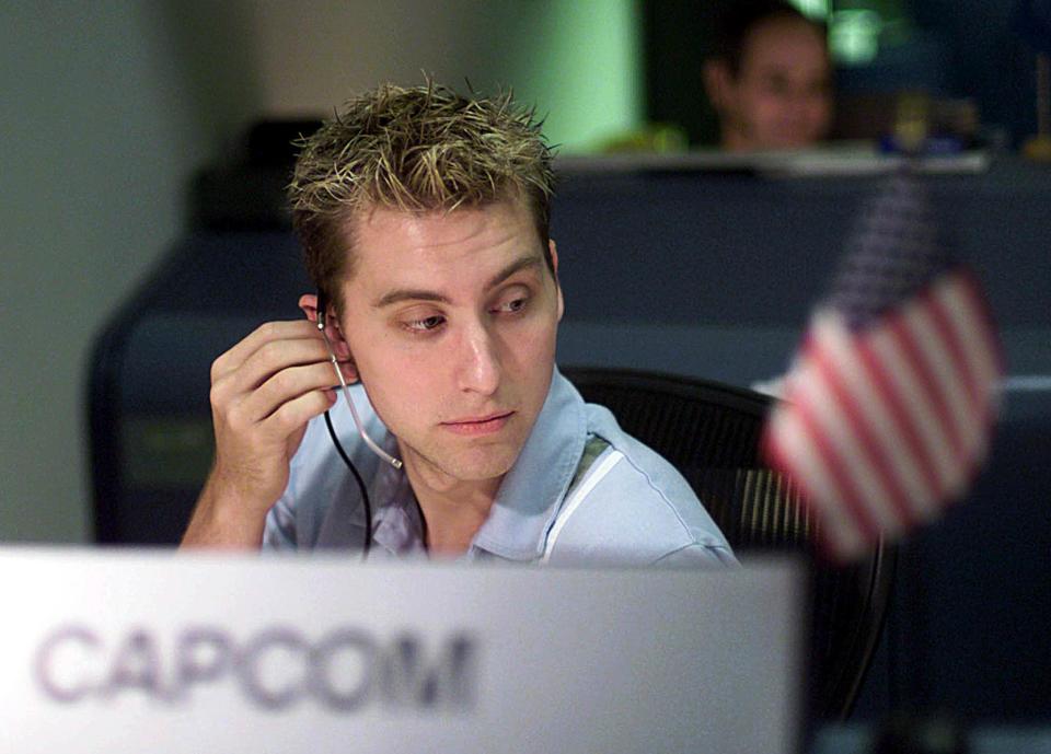 lance bass wears a headset while sitting down in an office chair. in front of him, blurry, is a sign with the word 