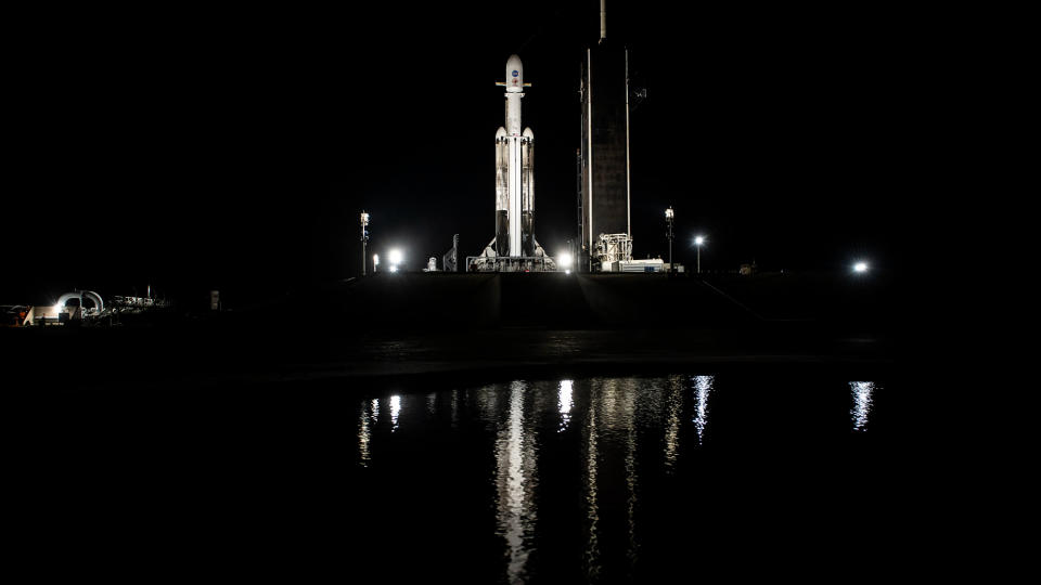 a white rocket sits on its launch pad at night, with a small lake in the foreground.