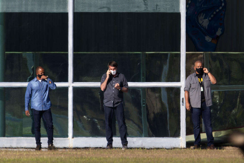 Brazil's President Jair Bolsonaro, center, talks on his phone while standing outside his official residence Alvorada Palace, in Brasilia, Brazil, Friday, July 10, 2020. Bolsonaro, 65, announced that he is infected with the new coronavirus on Tuesday and is using it to publicly extol hydroxychloroquine, the unproven malaria drug that he’s been promoting as a treatment for COVID-19, and now takes himself. (AP Photo/Eraldo Peres)