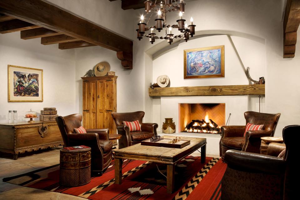 A western-style room with four brown, leather chairs around a coffee table with a chess board on top. Behind is a lit fire place on a white wall