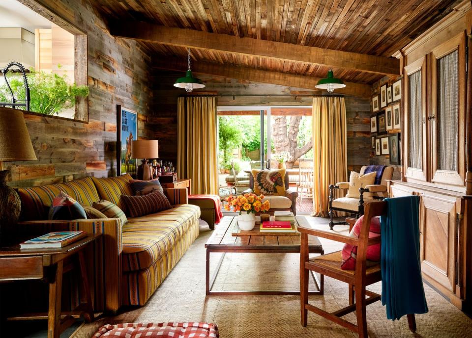 a family room with rustic wood ceiling and walls, sofa in a mustard fabric with stripes, cocktail table and end tables with lamps, three armchairs, built in wood cabinetry, framed artworks, sliding glass doors with curtains