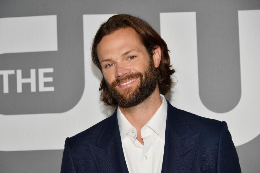 A man with long hair and a beard smiles while wearing a suit