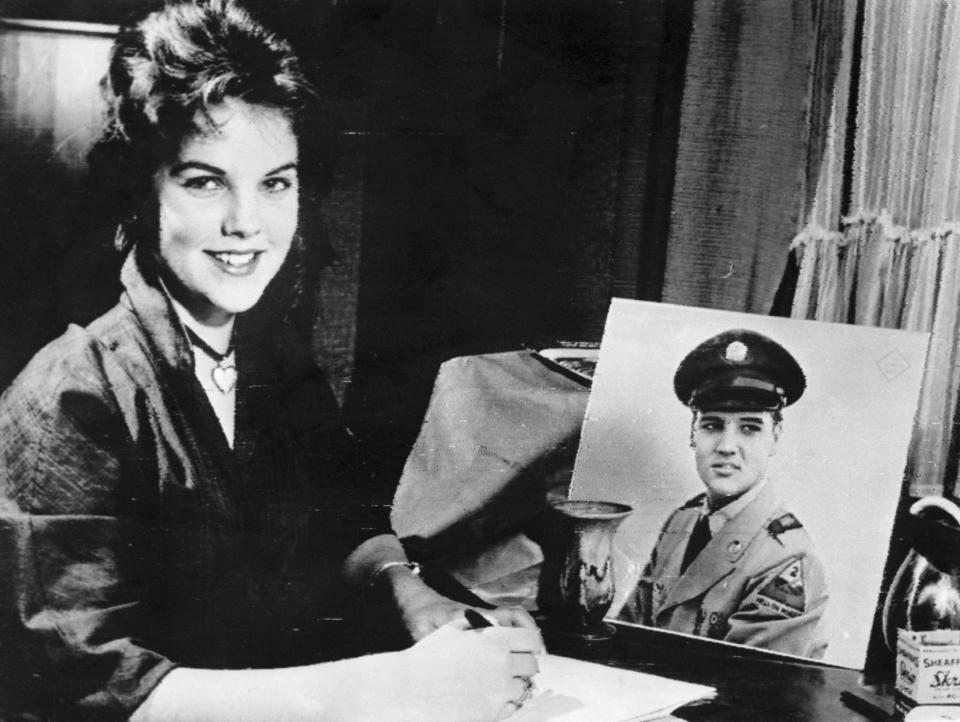 Priscilla smiling and sitting at a desk with a picture of Elvis in uniform