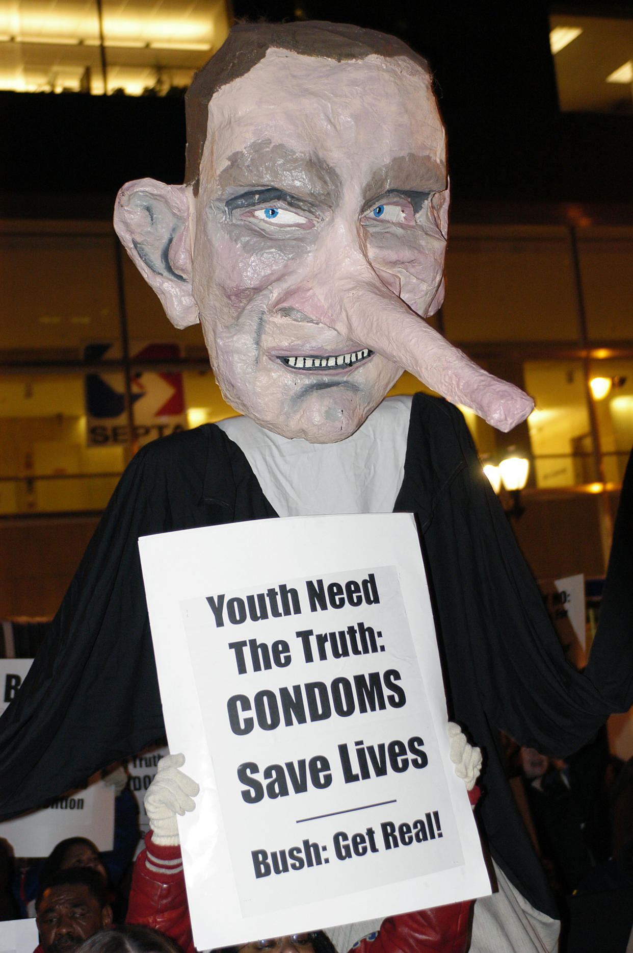 A giant puppet depicting President George W. Bush as Pinnochio at a 2004 protest holds a sign that reads: Youth need the truth: Condoms save lives. Bush: get real!