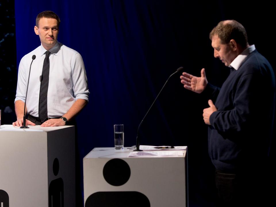 Alexei Navalny, standing in front of a podium, looks toward former Kremlin adviser Andrei Illarionov who is motioning with his hands as he speaks during a televised debate.