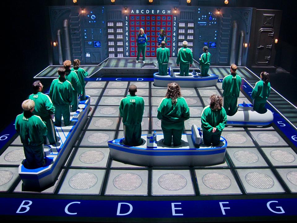 squid game players standing on a battleship game like set with a board and large, play ships