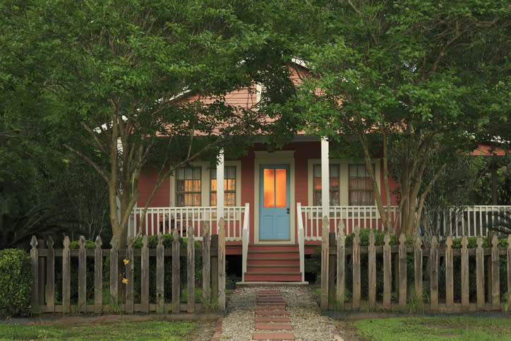 A quaint cottage-style house with a blue front door, lit warmly from the inside, surrounded by trees and a wooden picket fence in front