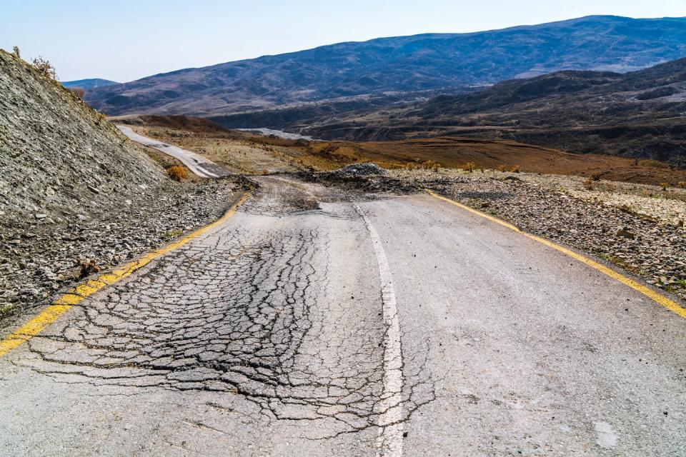 There is an isolated two-lane road, cracked and dilapidated, surrounded by hills and mountains.