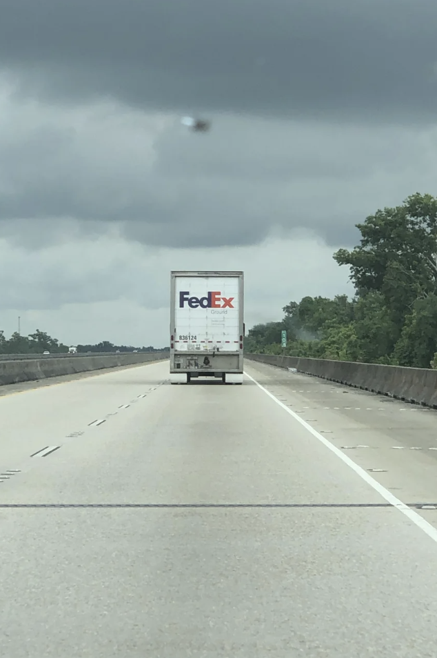 A FedEx truck is driving on an empty highway under a cloudy sky. Trees line the right side of the road