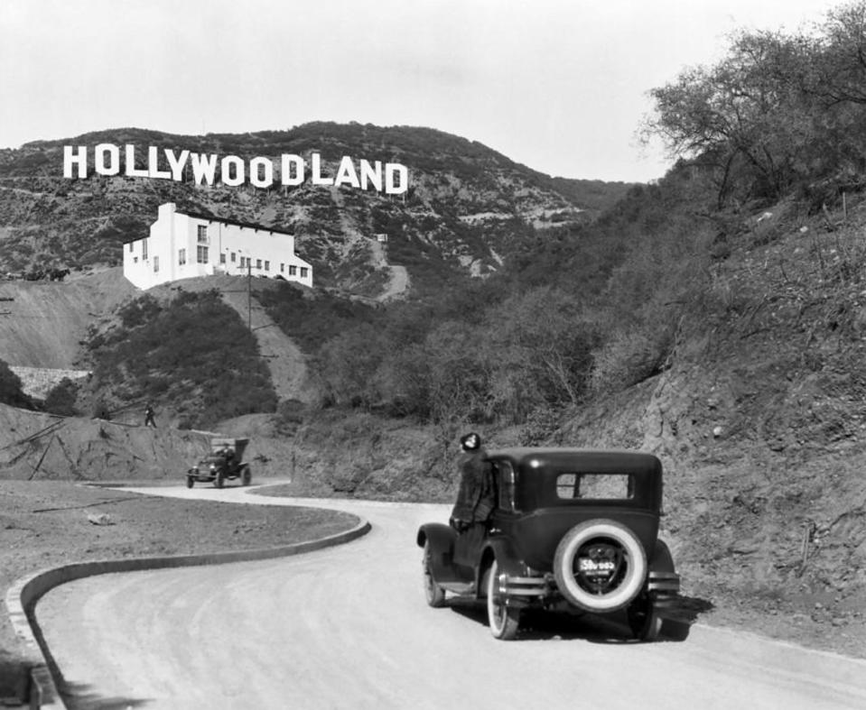 Driving on a winding road towards the original Hollywoodland sign on a hillside, featuring vintage cars from the early 20th century