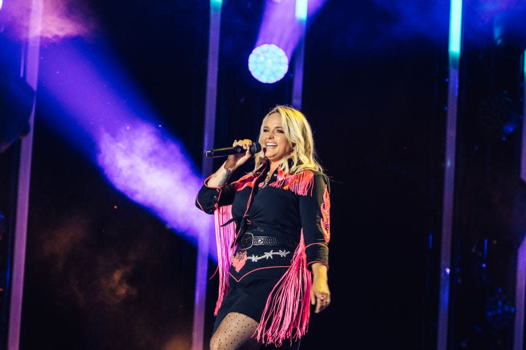 miranda lambert sings into a microphone she holds in one hand, she wears a dark outfit with hot pink fringe details