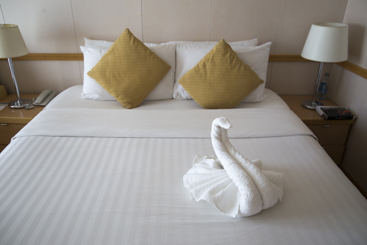 Bed in the bedroom cabin of a cruise ship, a towel animal swan in the foreground of a made-up bed, all white, two lamps and nightstand on both sides of the bed, and a wall