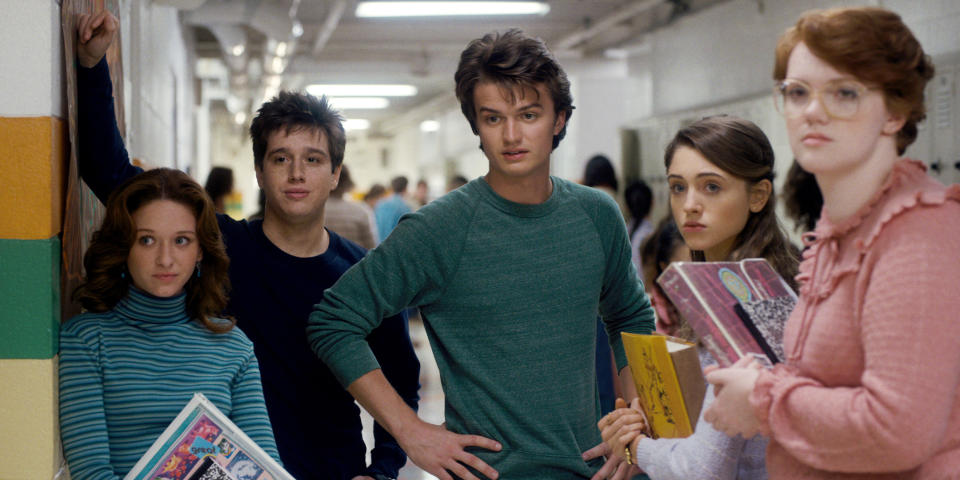 Five teens from "Stranger Things" with '80s school hallway backdrop, holding books, looking alarmed