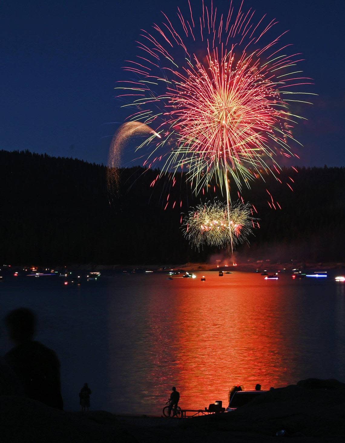 A cyclist and families watch from the shoreline at the 2022 Shaver Lake fireworks show Saturday, July 2, 2022 at Shaver Lake. A boat parade started at 6pm and the fireworks which began at dark were visible along the shore for thousands who came to celebrate.