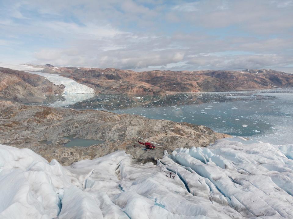 helicopter sits on rocky ledge in vast landscape with glacier sloping down to bare rock sloping down to open water full of icebergs