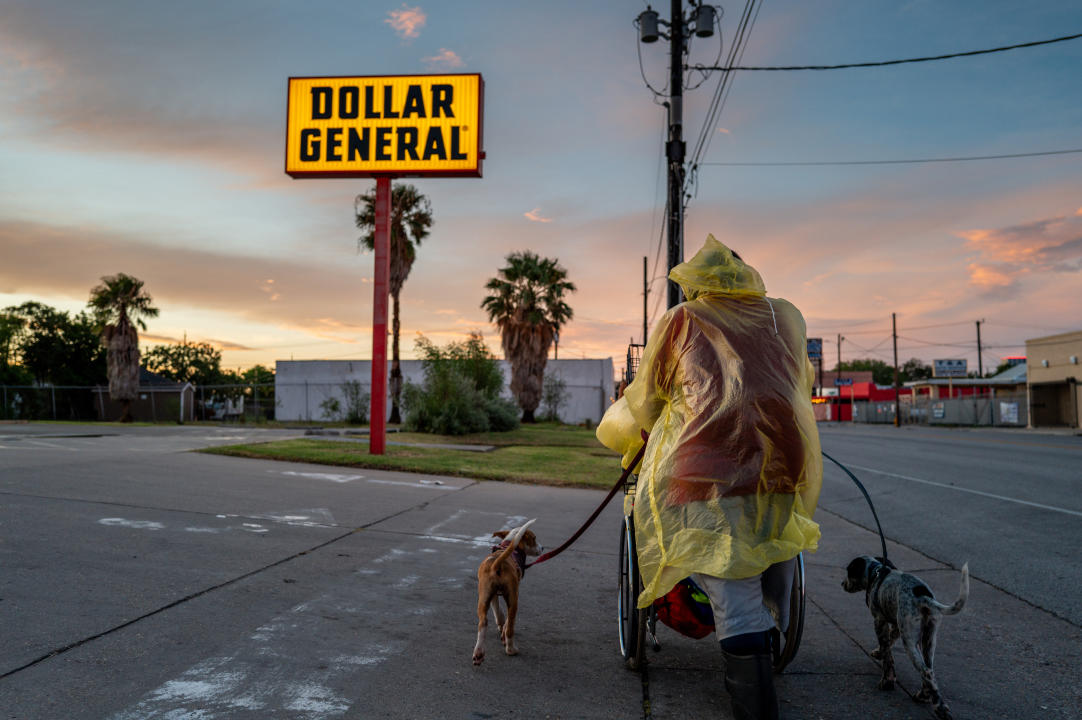 A person wearing a plastic poncho makes their way down an empty street near a Dollar General store with two dogs. 
