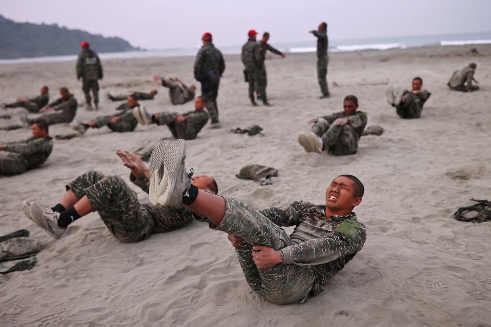 A man in a military uniform grimaces as he takes part in exercises with other men on a beach
