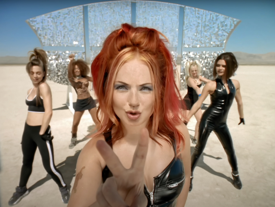 Geri Halliwell, Emma Bunton, Mel C, Mel B, and Victoria Beckham of the Spice Girls in futuristic outfits, performing a dance routine in a desert setting with a sparkly backdrop