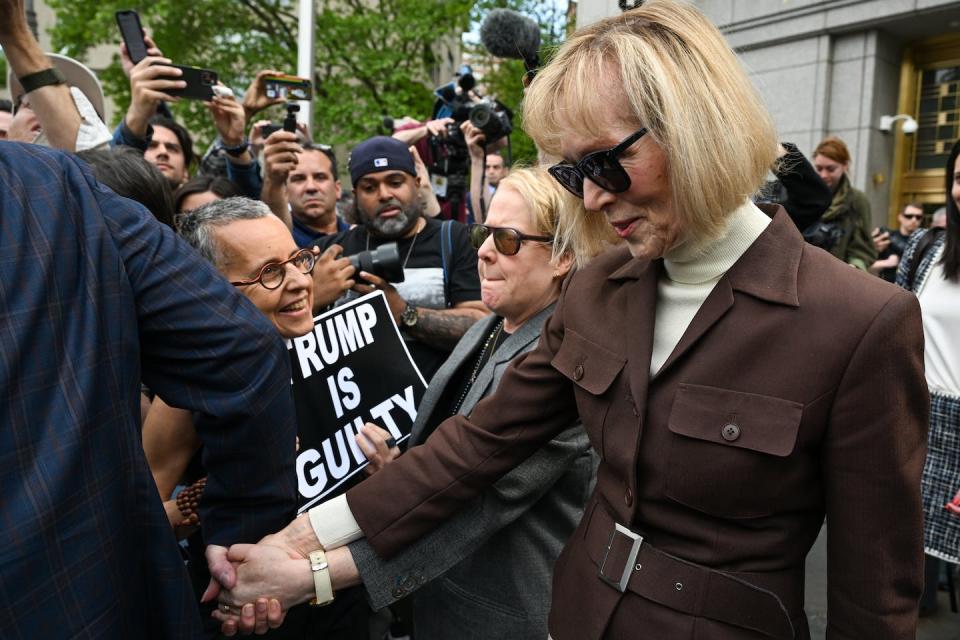 E. Jean Carroll walks through a crowd of people and photographers, including a person holding up a sign that says 'Trump is guilty.'