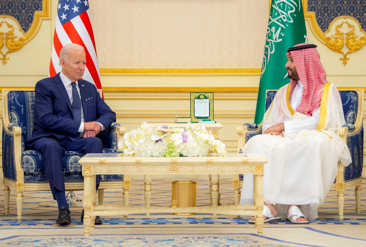 President Biden looking awkward, seated in a gilded armchair at a safe distance from Saudi Crown Prince Mohammed bin Salman, with their respective flags behind them. A low table set with a large dish of white flowers is set before them.