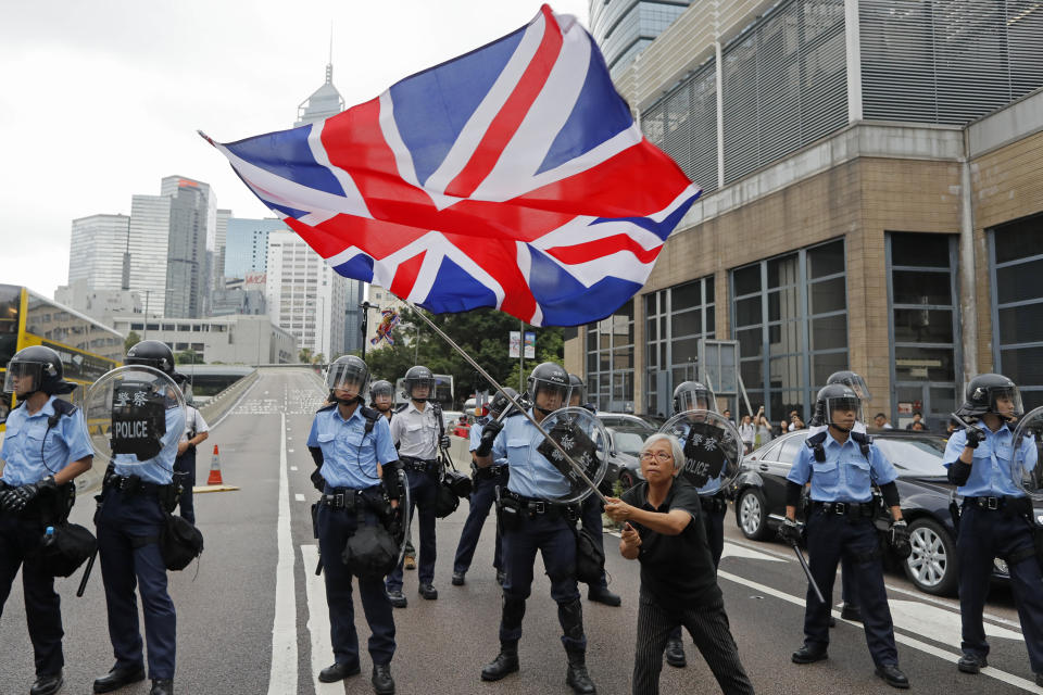 In this Wednesday, June 12, 2019, photo, a woman waves a British flag as policemen in anti-riot gear stand guard against the protesters on a closed-off road near the Legislative Council in Hong Kong. China promised that for 50 years after Britain gave up control of its last colony in 1997, this shimmering financial enclave would get to keep freedoms absent in the communist-ruled mainland that many here don't want to live without. (AP Photo/Kin Cheung)