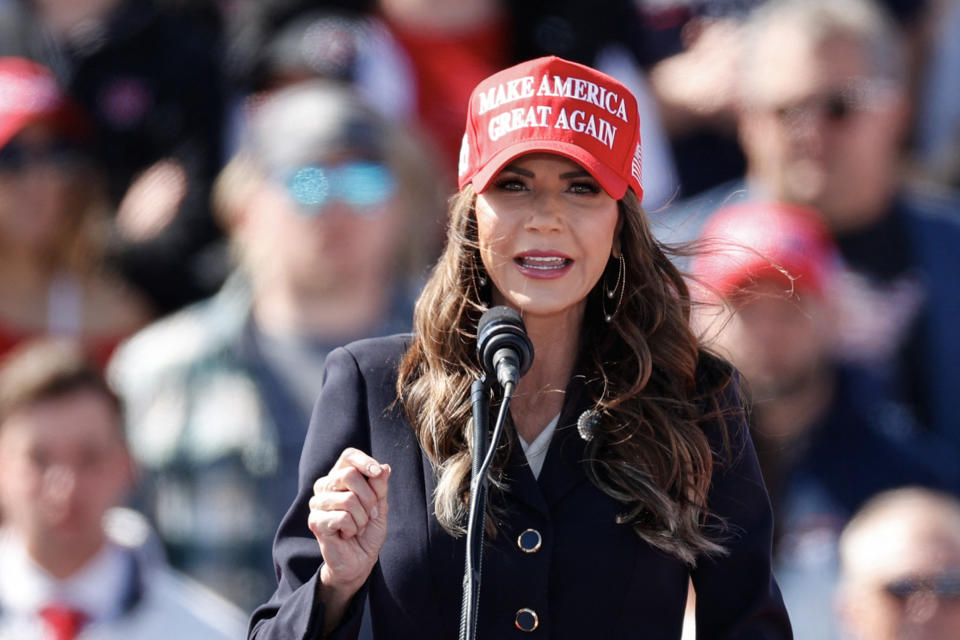 Woman speaking at a podium wearing a "Make America Great Again" hat