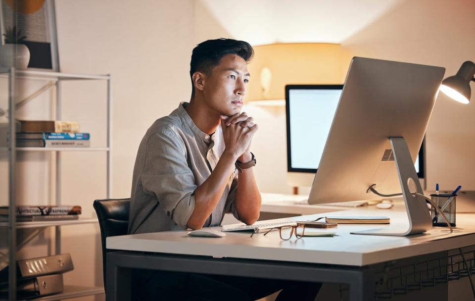 A person clasps their hands while looking at a desktop computer screen.