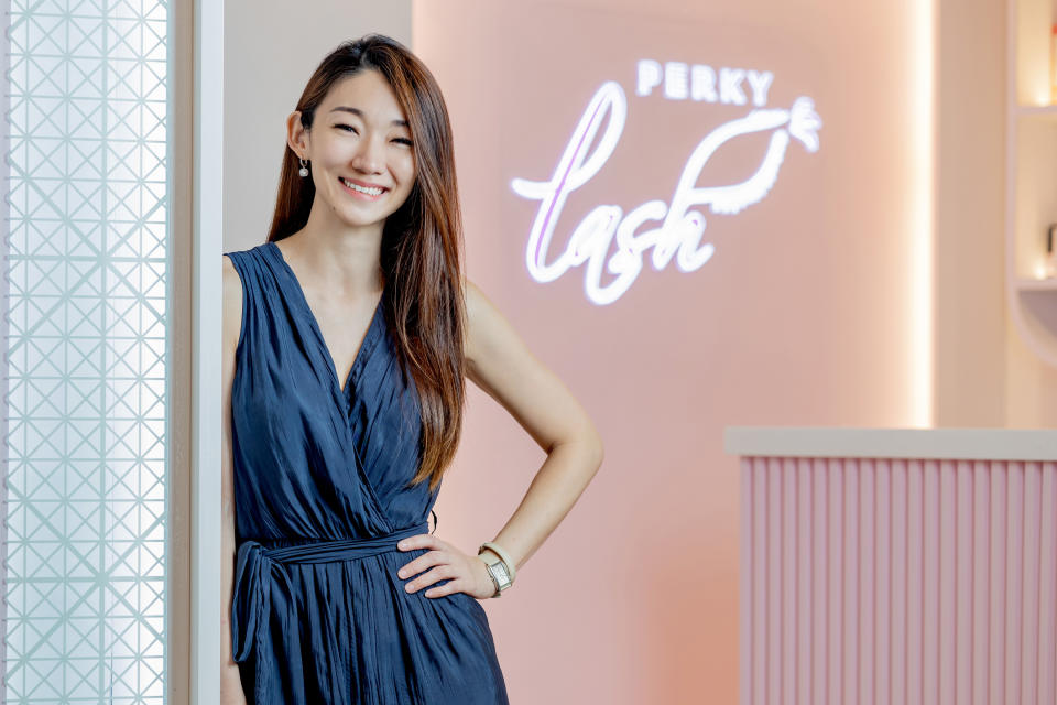 Jasmin Tay, the founder of Perky Lash, wearing a blue dress standing in front of a signboard in her shop. 