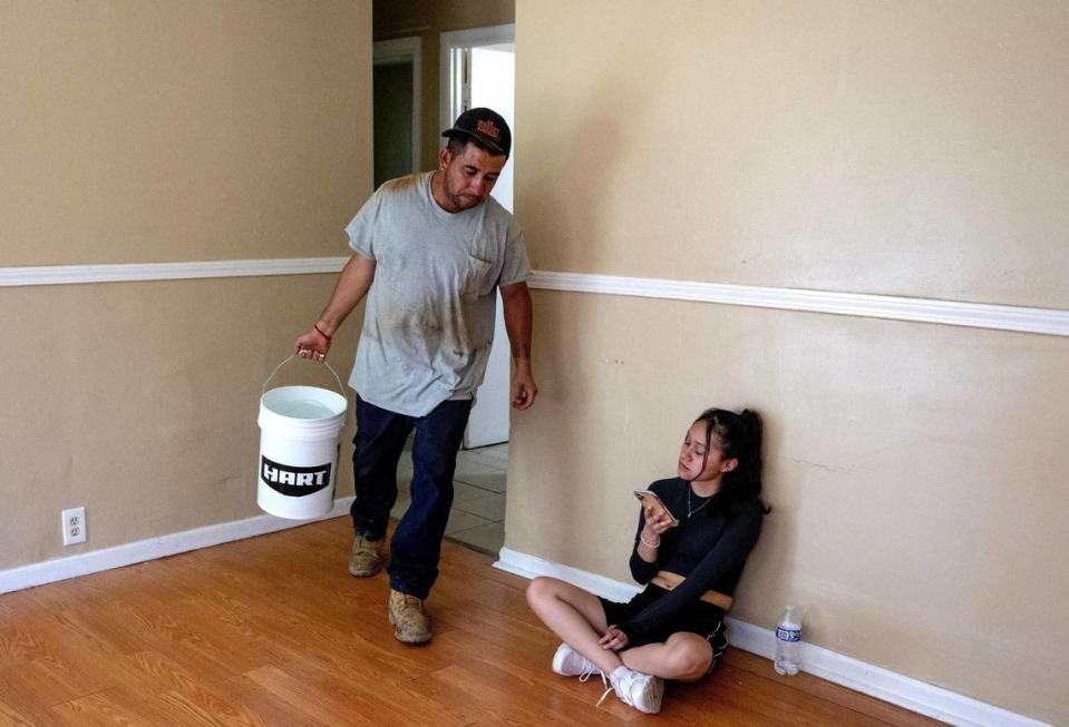 Alfonso López carries a bucket of water to the car as his daughter Geneva Martinez uses her phone on Friday, May 26, 2023, in Kansas City. Due to an outstanding balance left by a previous tenant, KC Water shut off water to López’s home in April. Since then, his family has had to get creative for their water needs including transporting old pop bottles and buckets full of water by car to their home and using bottled water to brush their teeth.