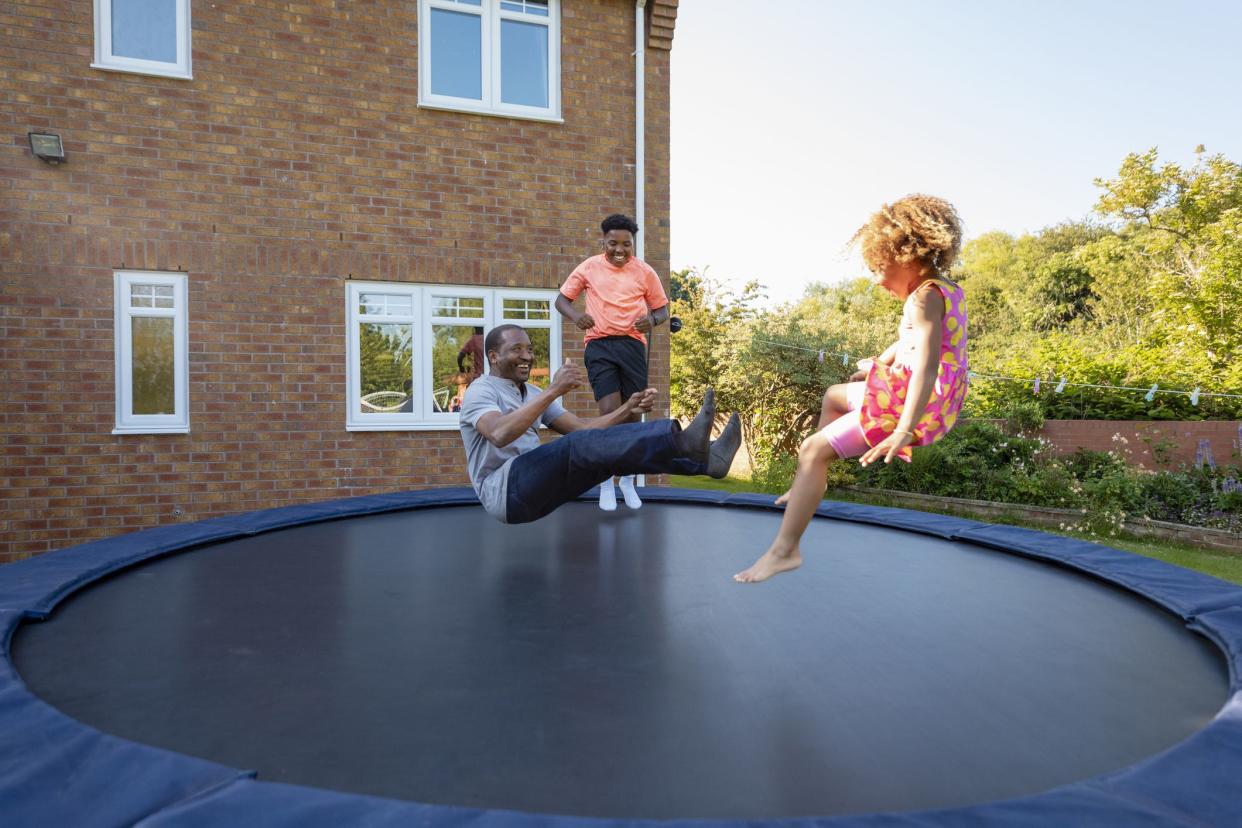 A young girl and her brother jumping on a trampoline outdoors in the back garden at home with their grandfather. They are all trying to do a seat drop jump.