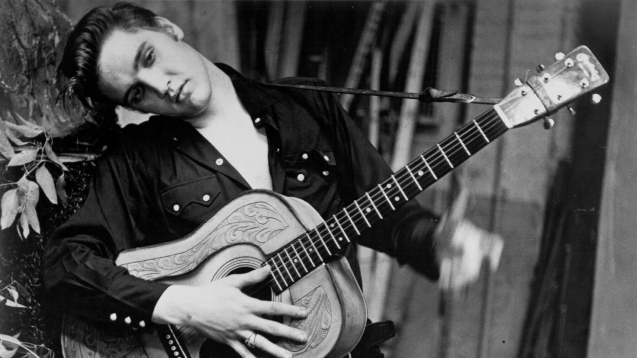 elvis presley leans against a wall as he holds an acoustic guitar