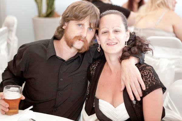 Dave holds a beer at a formal event with his arm around Allira. 