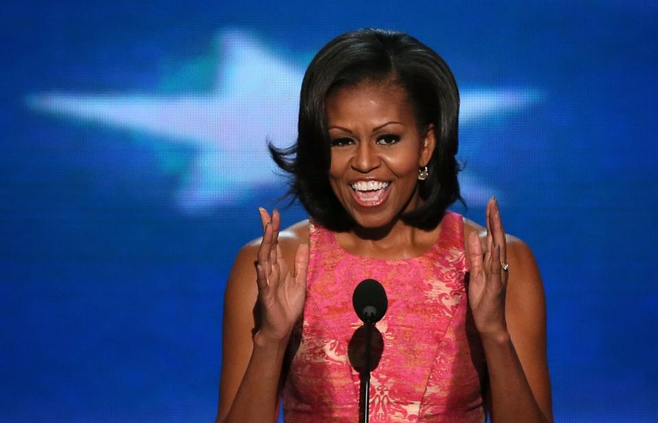 michelle obama, wearing a pink dress, speaking into a camera, with a blue and white backdrop behind her