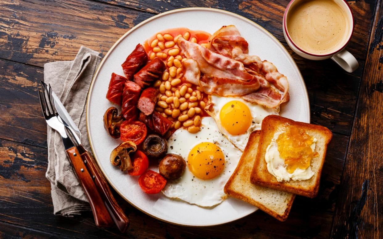 Full English breakfast with fried eggs, sausages, bacon, beans, toasts and coffee on a wooden background - The Picture Pantry/Getty Images