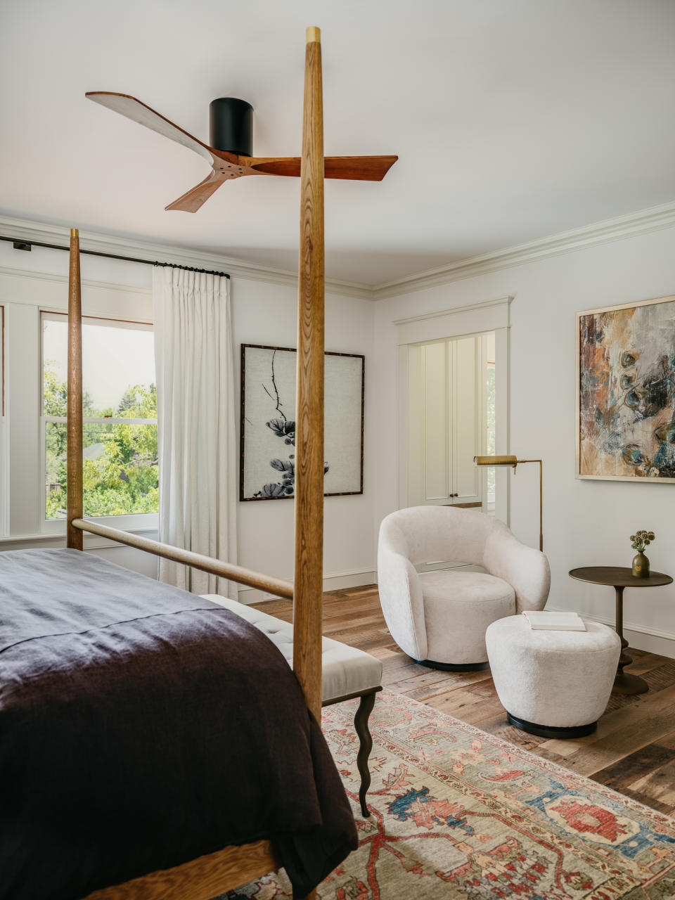 Wooden four-poster bed with dark throw, white armchair and footstool, antique rug and wall art