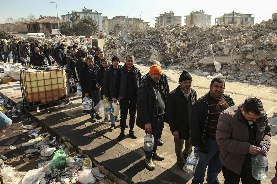 People line up for water by collapsed buildings in Kahramanmaras, southern Turkey, Sunday, Feb. 12, 2023. Six days after earthquakes in Syria and Turkey killed tens of thousands, sorrow and disbelief are turning to anger and tension over a sense that there has been an ineffective, unfair and disproportionate response to the historic disaster. (AP Photo/Emrah Gurel)