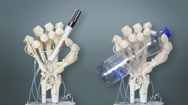 Image of two all-in-one 3D-printed robotic hands, one holding a marker pen, the other gripping an empty clear plastic water bottle. Both are stood on plinths in front of a dark grey background.