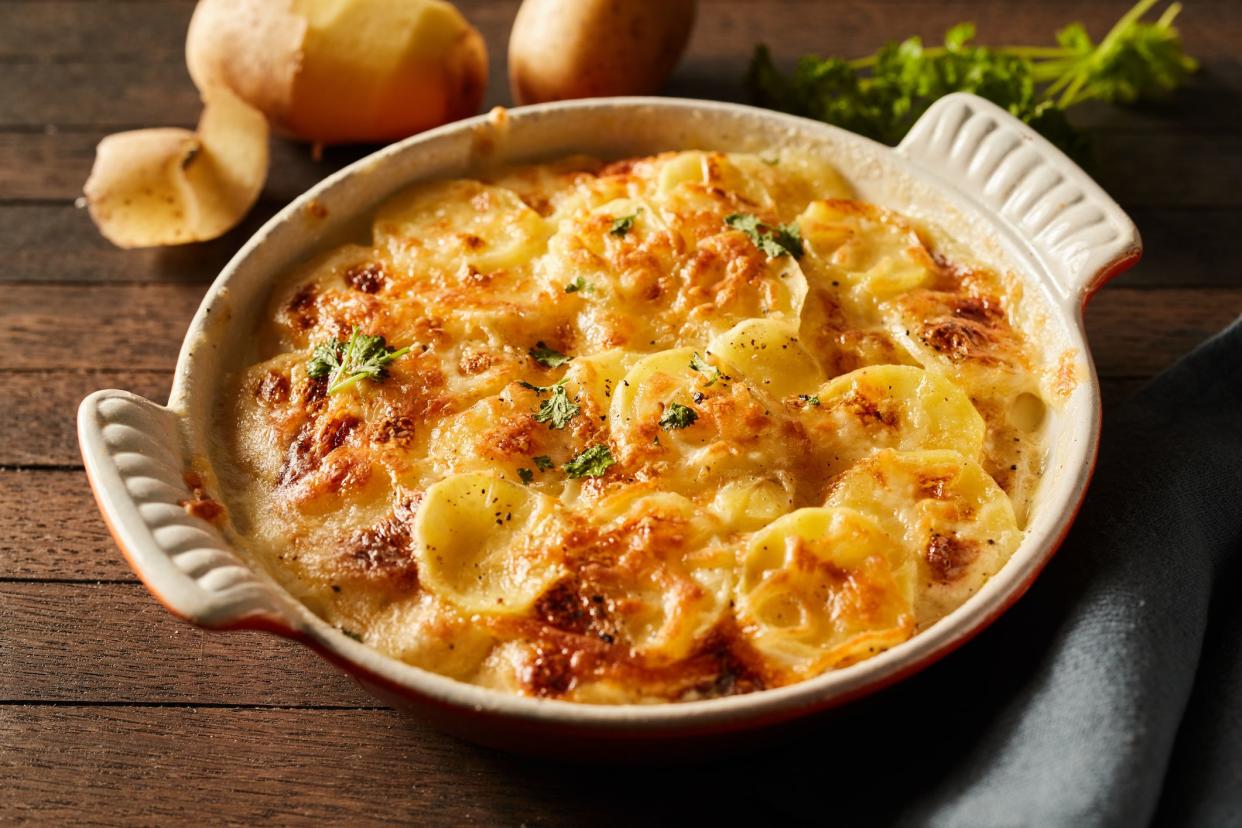 High angle of scalloped gratin potatoes topped with grated cheese and herbs in ceramic baking dish on table with towel and branches of greens