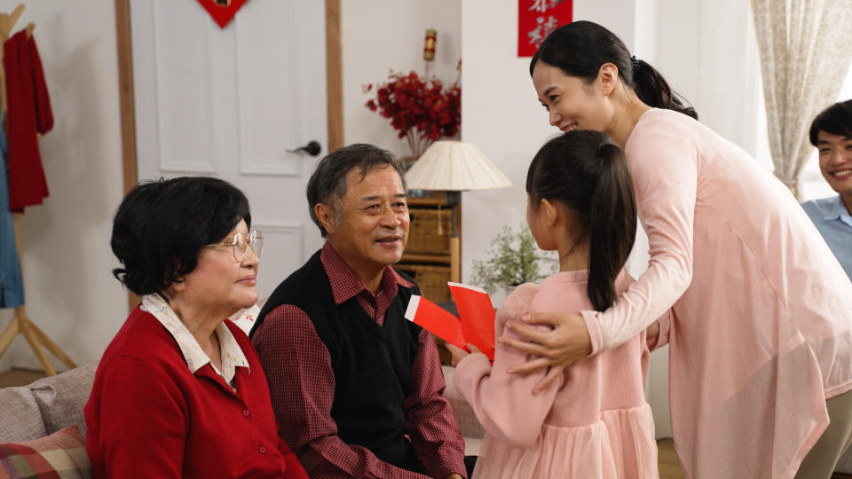 happy asian elderly couple grandmother and grandfather telling grandchild to come, giving red envelope and wishing her happy chinese new year with hand gestures in the living room at home